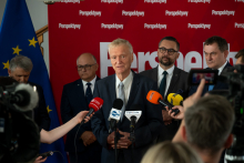An older man speaks during a press conference, while journalists record the event and several people stand behind him.