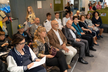 A group of people sits on chairs in a room taking part in an event. Some individuals hold documents or phones in their hands. In the background, more people stand, and coat racks with bags and gray walls are visible.