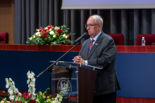  One person stands at a podium and speaks. A floral arrangement is placed in front of the podium, with the lecture hall stage in the background.