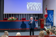 One person stands in front of the stage and speaks into a microphone. A screen behind shows a black-and-white group photograph and elements of a lecture hall.