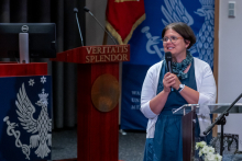  One person stands at a podium speaking into a microphone. Elements of the stage and the university emblem are visible in the background.