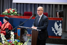 A man in a suit stands at a lectern with a microphone, resting hands on the podium while speaking. In the background there is a person in an academic gown and cap, as well as stage elements with the university emblem and floral decorations.
