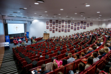 Large auditorium filled with several dozen graduates and guests seated in rows. Many graduates wear academic caps. A stage with seated officials is visible at the front, along with a screen displaying the Medical University of Warsaw logo.