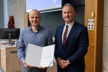 Two men stand next to each other, the man on the left holds an open diploma in his hands.