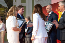 A woman and a man exchange a handshake on stage during an awards ceremony, while other participants hold framed certificates.