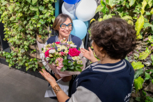 Two women stand facing each other. The woman on the left holds a large bouquet of flowers and a card, while the woman on the right gestures with both hands during the conversation. A green plant wall and white-and-blue balloons are in the background.