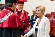 A person in a light jacket shakes hands with a person wearing a red and black academic gown with a decorative chain. Another person in a gown stands nearby holding an open folder with a document. Red auditorium seats and additional participants are visible in the background.