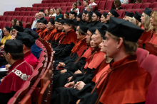 A group of people in gowns and mortarboards are seated in an auditorium on red chairs.