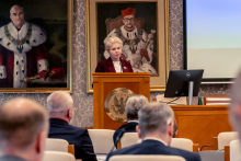 One person stands at a lectern and addresses conference participants. Portraits and a presentation screen are visible in the background. Several seated participants are in front of the lectern.