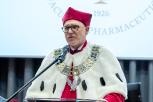 One official speaks at a lectern during a ceremony, wearing academic robes and a ceremonial chain. A microphone and university backdrop are visible.