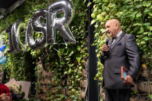 A man stands by a green plant wall and speaks to the audience. The man holds a microphone in one hand and a book or notebook in the other. Silver balloons forming the word “CSR” are placed beside him.