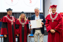 Two people and one additional person wearing red and black academic gowns stand beside a person in a jacket. The person in the jacket holds an open folder with a document and presents it. In the background, a screen with graphics and elements of the lecture hall can be seen.