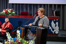 A person speaks at a podium during a formal event in a conference hall; national symbols and floral decorations are visible in the background, and on the left side a representative in ceremonial attire is seated.