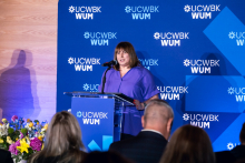 One person stands at a podium and speaks to the event participants. A UCWBK WUM logo backdrop is visible behind, with flower arrangements placed beside the stage.
