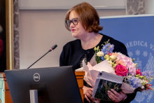 One person stands at a lectern with a microphone, holding a bouquet of flowers and documents while speaking. A monitor and a university banner are visible nearby.