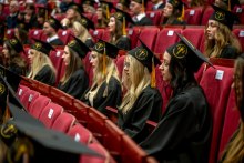 Group of graduates sits in rows in an auditorium, wearing academic caps and gowns.