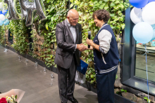 A woman and a man stand facing each other and shake hands. The woman holds a microphone in her hand, and the man holds a folder and a bag. A green plant wall and decorative balloons can be seen in the background.