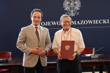 Dwaj mężczyźni stoją przed tłem z napisem „Wojewoda Mazowiecki”. Mężczyzna po prawej trzyma czerwoną teczkę z godłem.
