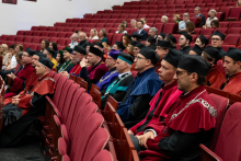 A group of people in gowns and mortarboards are seated in an auditorium on red chairs.