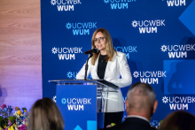 One person stands at a podium and speaks to the event participants. A UCWBK WUM logo backdrop is visible behind, and participants can be seen in the foreground.
