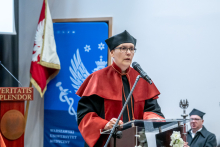 One official speaks at a lectern with a microphone. A flag and university banner are visible in the background, and another person stands behind.