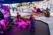 Journalists and camera operators film a medical simulation exercise in front of a large screen showing a virtual road traffic accident scene with a damaged car.
