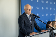 Jedna osoba przemawia do mikrofonu przy mównicy. W tle znajduje się ścianka z powtarzającym się napisem Warszawski Uniwersytet Medyczny oraz częściowo widoczne osoby siedzące obok.