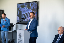 Dwie osoby stoją przy mównicy i pulpicie, jedna przemawia, druga trzyma dokumenty. Obok siedzi jedna osoba. W tle widoczny ekran z prezentacją budynku.