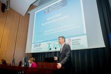 Jedna osoba prowadzi wykład stojąc przy mównicy. W tle wyświetlana jest prezentacja pt. „Head & Neck Reconstructive Surgery”. Przy stole obok siedzi druga osoba obsługująca sprzęt komputerowy.