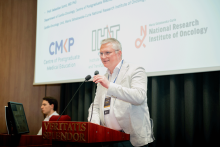 Jedna osoba przemawia do mikrofonu przy mównicy na sali wykładowej. Za nią wyświetlana jest prezentacja z logotypami CMKP, IITD i Narodowego Instytutu Onkologii. W tle widoczna jest druga osoba siedząca przy komputerze.
