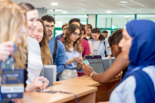 Kilka osób stoi w kolejce przy stanowisku rejestracyjnym. Pracownicy obsługują uczestników i przekazują identyfikatory. Na ladzie znajdują się materiały informacyjne oraz urządzenia do obsługi rejestracji.