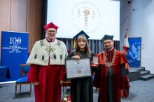 Trzy osoby ubrane w togi akademickie stoją na scenie. Jedna osoba trzyma dyplom i medal, a po obu stronach stoją przedstawiciele władz uczelni.