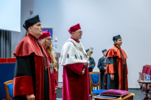 Cztery osoby w togach akademickich stoją na scenie podczas oficjalnej części uroczystości. W tle elementy sali i zgromadzeni uczestnicy.