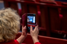 Jedna osoba trzyma telefon i wykonuje zdjęcie wnętrza sali wykładowej. Na ekranie telefonu widoczna jest scena z prezentacją.
