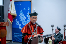 Mężczyzna, dziekan WF, w stroju akademickim przemawia przy mównicy podczas ceremonii WUM. W tle widoczne symbole uczelni i osoby siedzące na scenie.