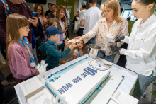 Przy stole z dużym tortem z napisem „10 lat CSR WUM” stoi kilka osób. Dzieci podają sobie kawałek tortu na talerzyku, a dorosłe osoby porcjują kolejne kawałki przy użyciu łopatek i talerzy. Na stole znajdują się plastikowe sztućce, serwetki i metalowe wiadro. W tle stoją inni uczestnicy wydarzenia obserwujący sytuację.