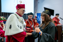 Młoda kobieta odbiera dyplom lub wyróżnienie od przedstawiciela władz uczelni na scenie. W tle stoją inne osoby uczestniczące w ceremonii.