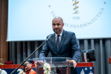 Elegancko ubrany mężczyzna przemawia przy mównicy na tle ekranu z logo Wydziału Farmaceutycznego.