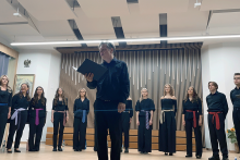 Grupa 12 osób stoi na scenie. Jedna osoba stoi na przedzie, trzyma w rękach teczkę.