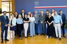 Grupa osób pozuje do zdjęcia. Za nimi widać niebieską ściankę z herbem Polski, pasem w kolorze biało-czerwonym oraz napisem: "Permanent Mission of..."
