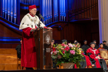 A man in a ceremonial rector’s robe speaks at a lectern. Other members of the academic community are seated behind him.