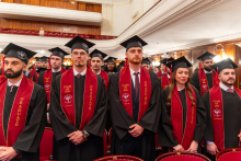 A group of male and female graduates in academic gowns and caps stand in an auditorium during the ceremony.