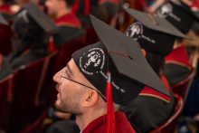A man wearing glasses and a graduation cap sits in an auditorium and looks toward the stage.