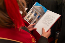A graduate holds an open commemorative book in their hands and reads its contents while seated among other participants of the ceremony.