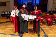 Three men pose for a photograph on stage. The graduate in the center holds a diploma folder and a framed certificate in his hands.