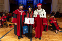 A woman in a graduation gown and cap poses with two men in ceremonial academic robes. She holds a diploma folder in one hand and a university gift bag in the other.