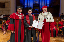 A man in a graduation gown and cap poses with two university officials in ceremonial robes while holding an open diploma folder in his hands.