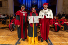 A woman in a graduation gown and cap poses with two men in ceremonial academic robes. She holds an open diploma folder in her hands.