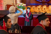 A woman in a graduation gown and cap poses for a photograph while holding a diploma and a commemorative paddle. In the foreground, another woman is taking her picture.