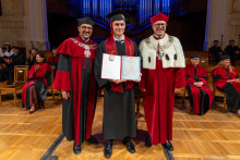A man in a graduation gown and cap stands between two university officials in ceremonial robes and holds an open diploma folder in his hands.